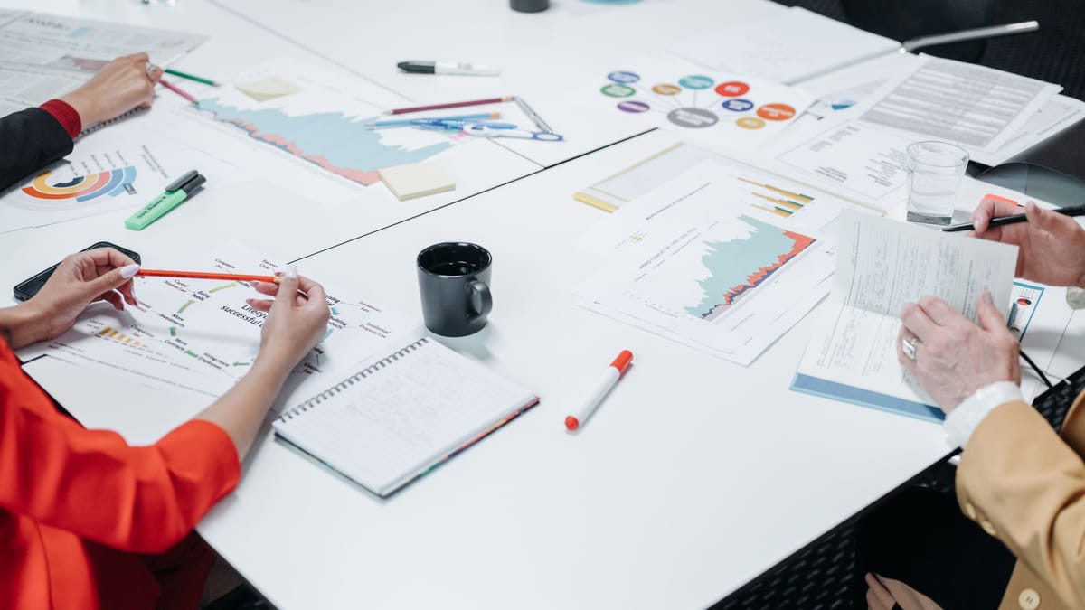 Paperwork, charts, markers, and office supplies spread across a white desk.