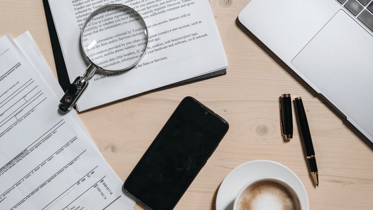 Documents, a magnifying glass, pens, a phone, and a laptop on a desk.