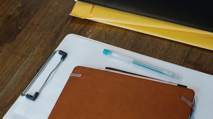 A clipboard, notebook, folder, and pen arranged on a wooden desk.